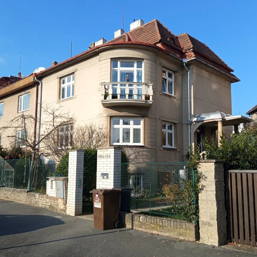 Sunny day with beige two-story house, red tiled roof, garden, and street entrance