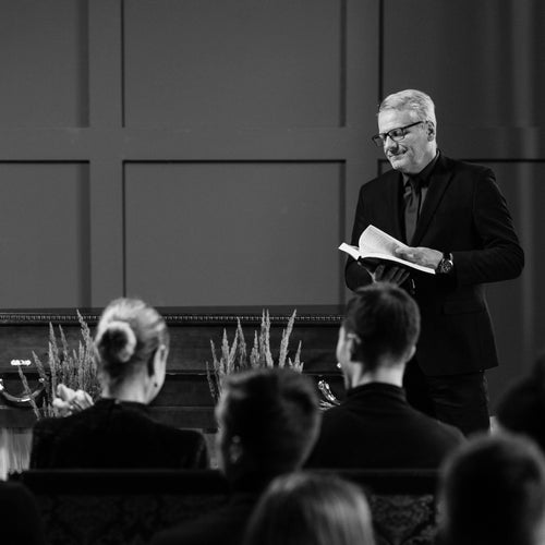 Man speaking at a funeral gathering with mourners seated, black and white photo