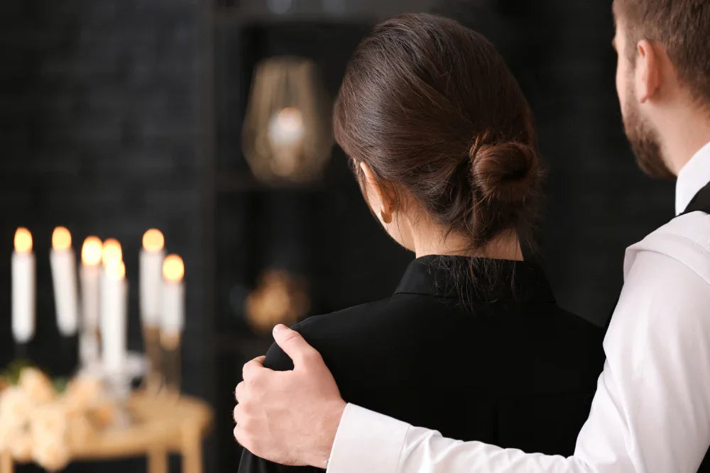 Two people in mourning embrace at a funeral reception with candles in the background.